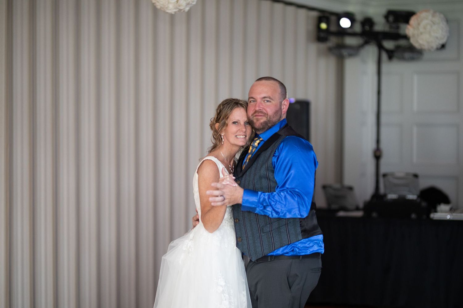 Bride and groom share first dance at wedding reception, groom in blue shirt and vest, bride in white gown.