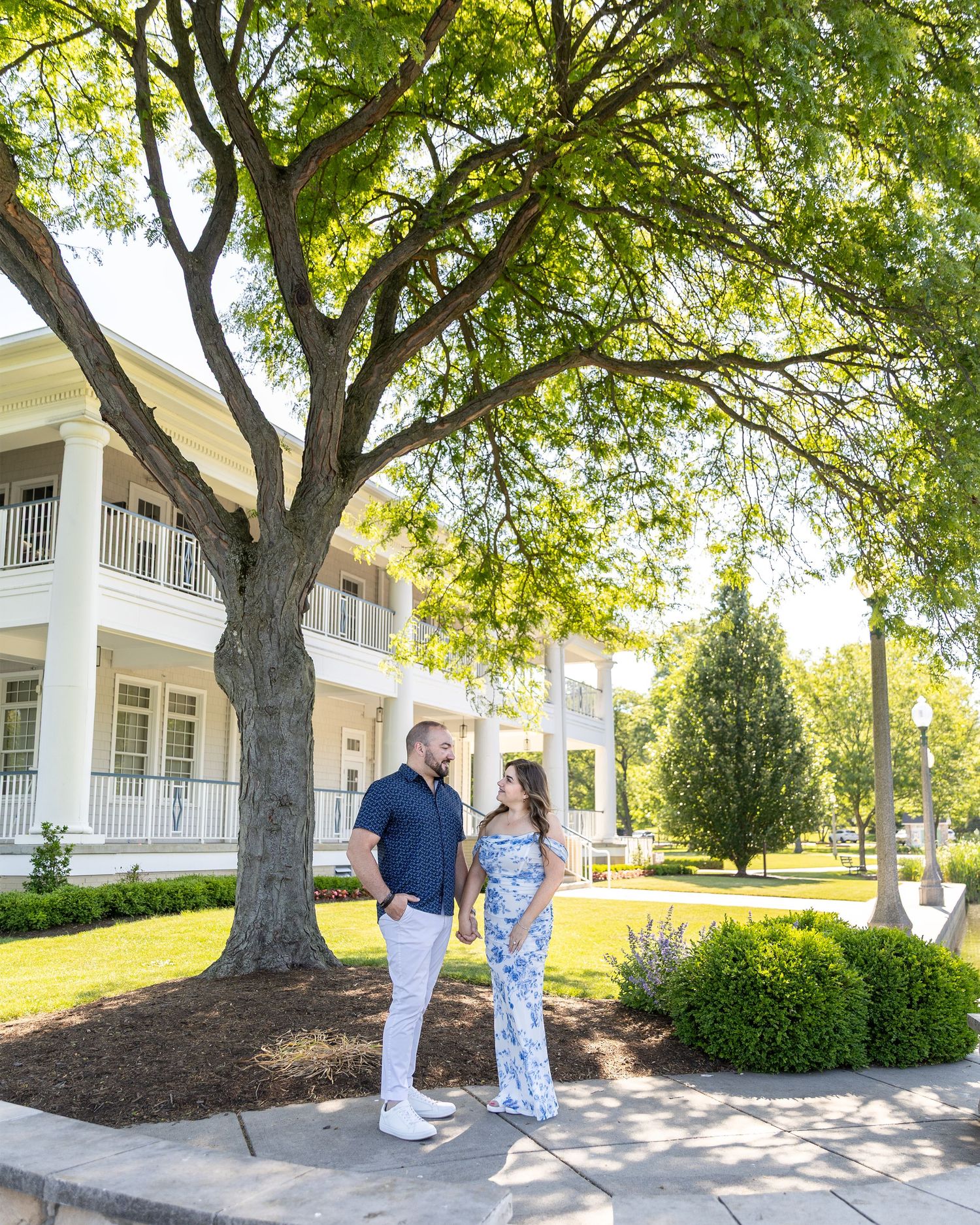Couple standing under a large tree in front of a white Southern-style mansion on a sunny day.