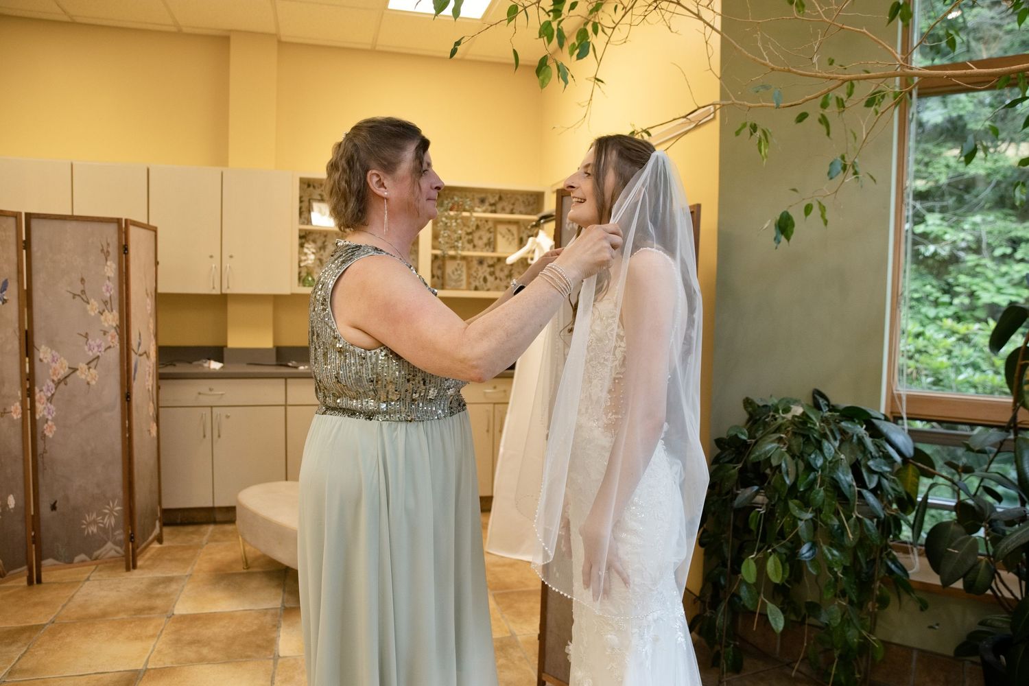A bridesmaid in blue adjusts a bride's veil in a sunlit room before the wedding ceremony.