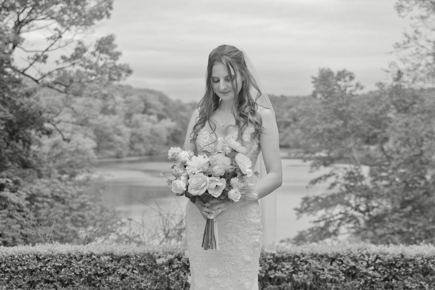 Elegant bride in fitted lace wedding gown holds a bouquet of roses in a black and white outdoor portrait.