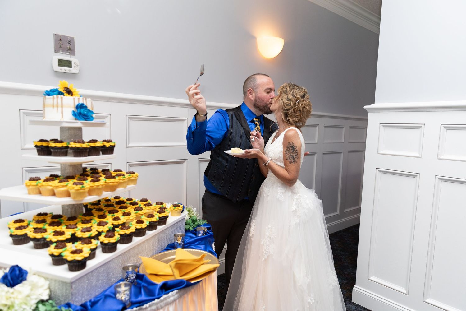 Bride and groom sharing a kiss while cutting their wedding cake surrounded by sunflower cupcakes at their reception.