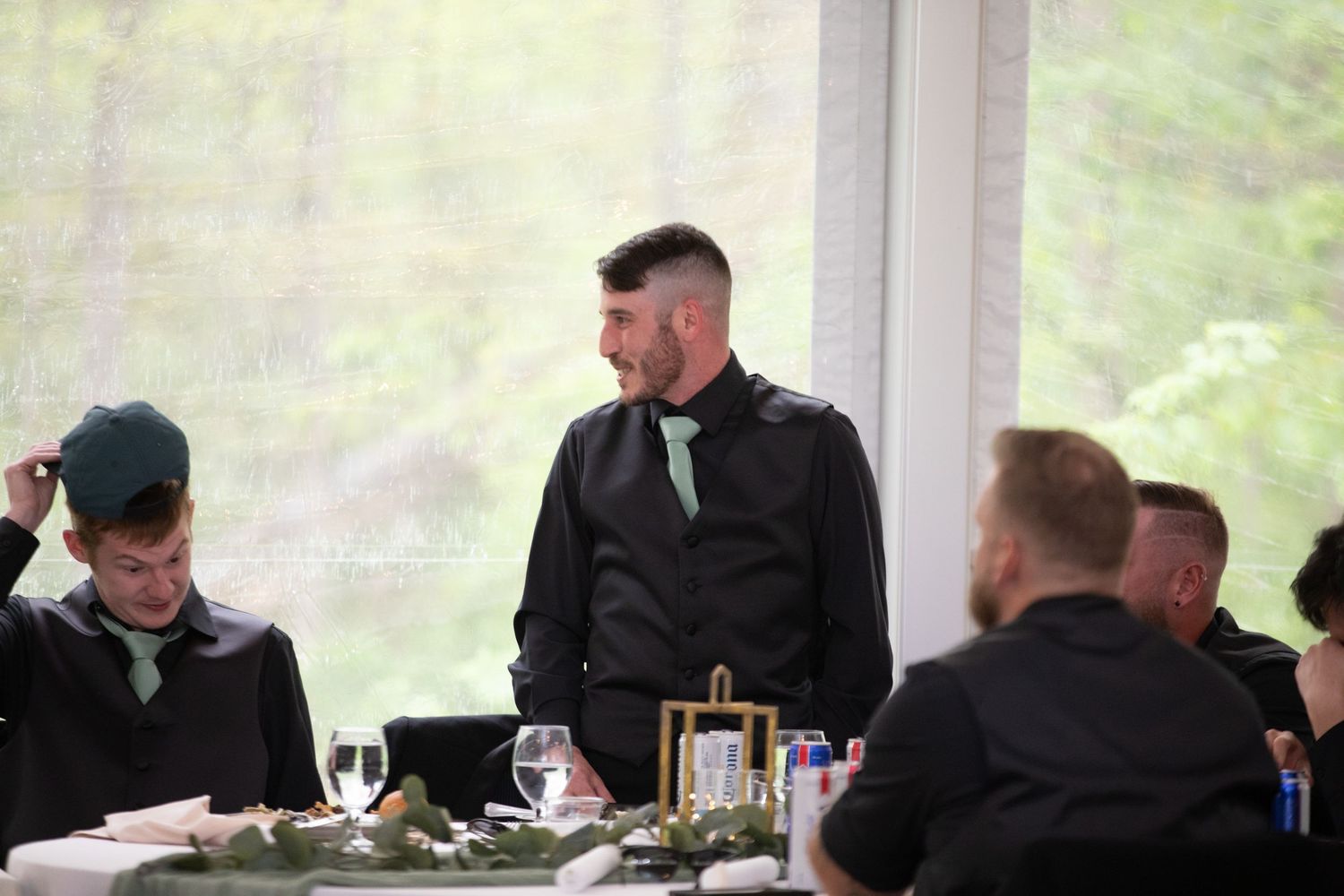 Men in black suits with green ties gathered at an elegant wedding reception table near large windows.