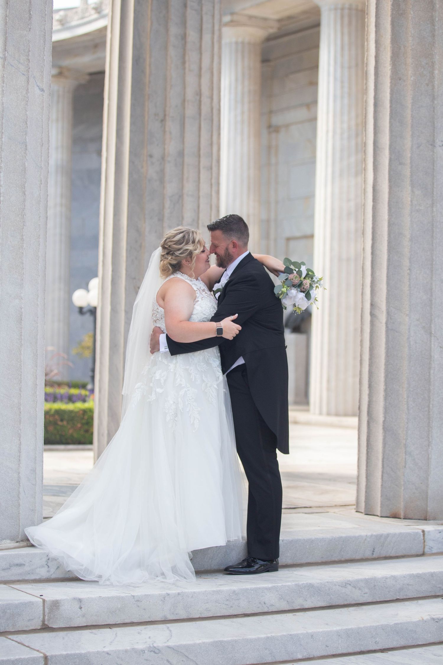 A bride and groom share a romantic moment between classical stone columns.
