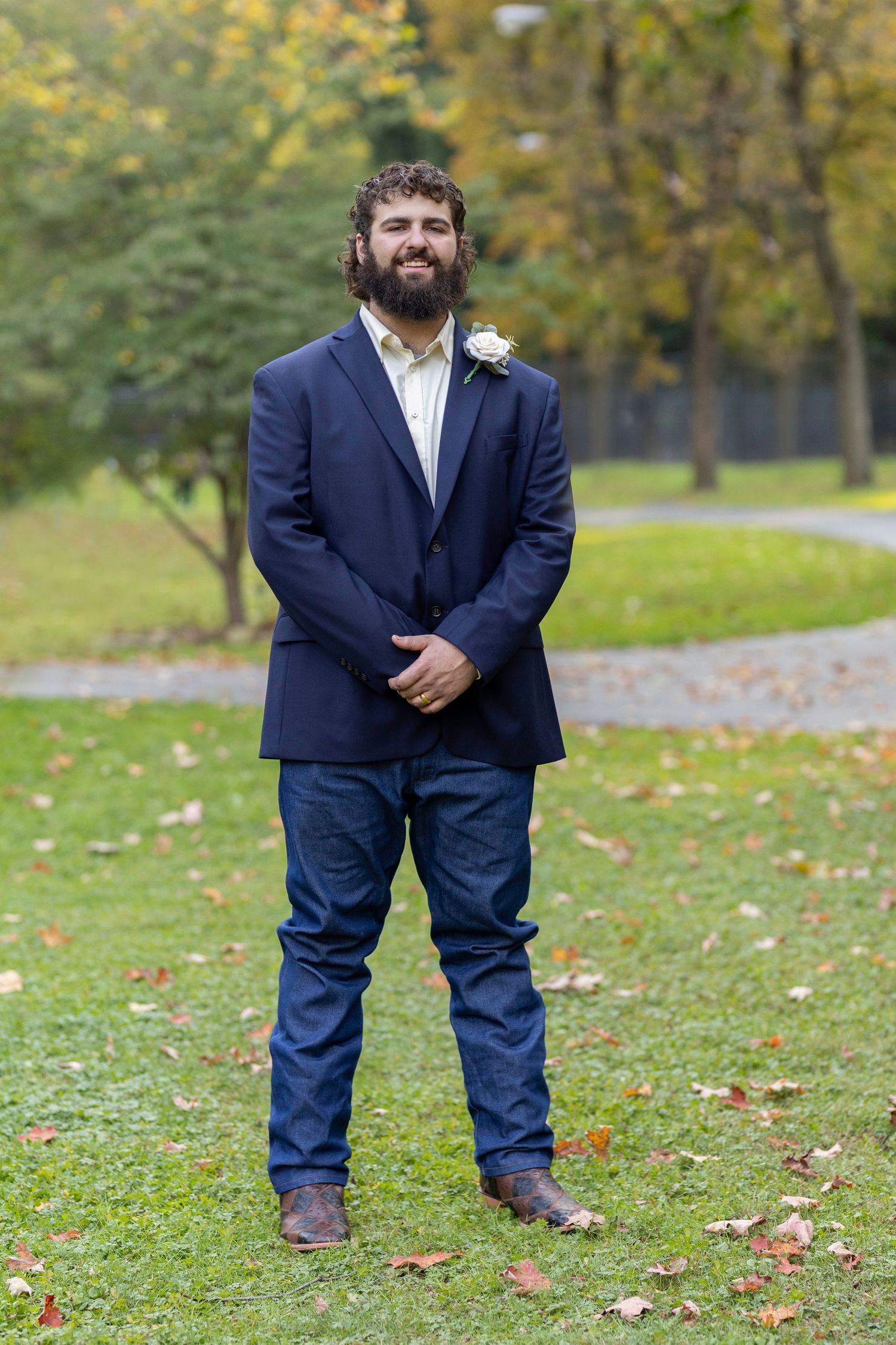 A groom in a navy suit and white boutonniere stands on grass with autumn trees in background.