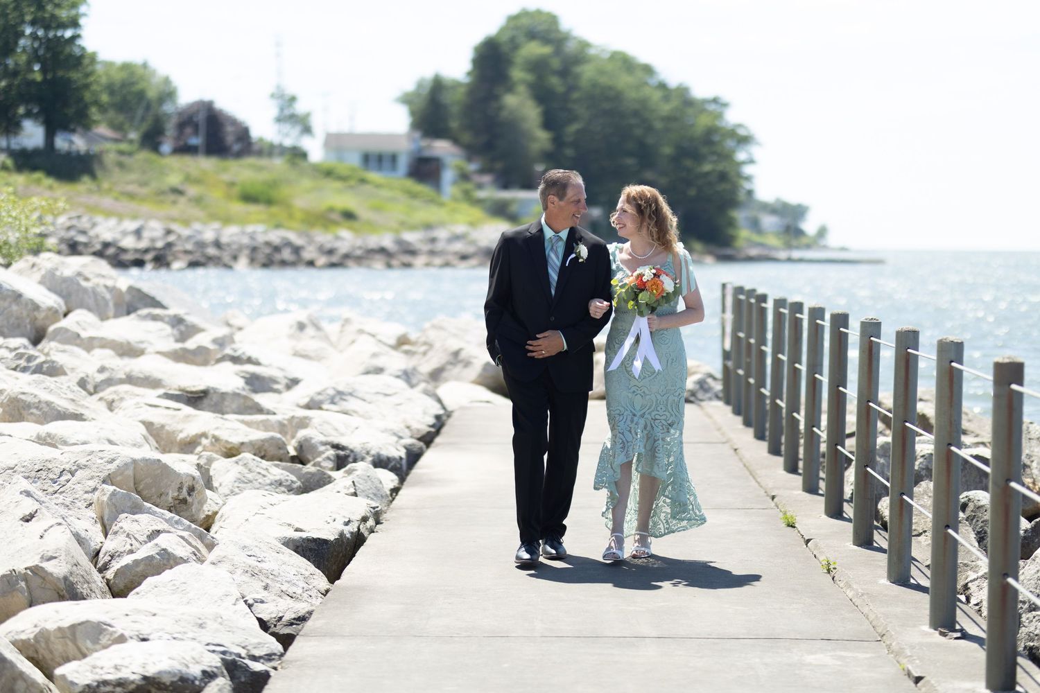 Couple strolling along a rocky shoreline with wooden fence and water views.