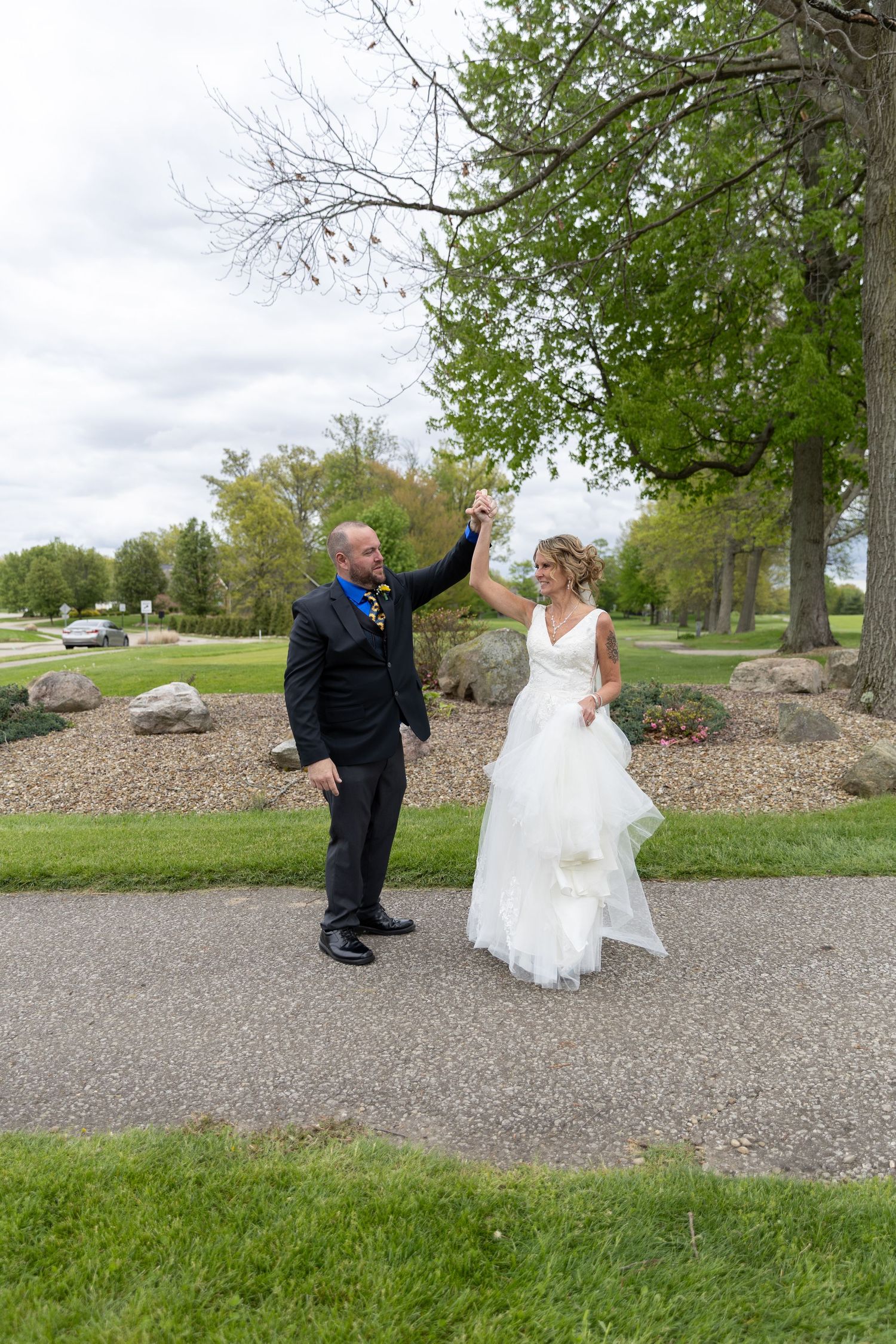 Bride and groom dancing outdoors on a path, groom in black suit with blue tie, bride in white layered gown, lush green trees behind.