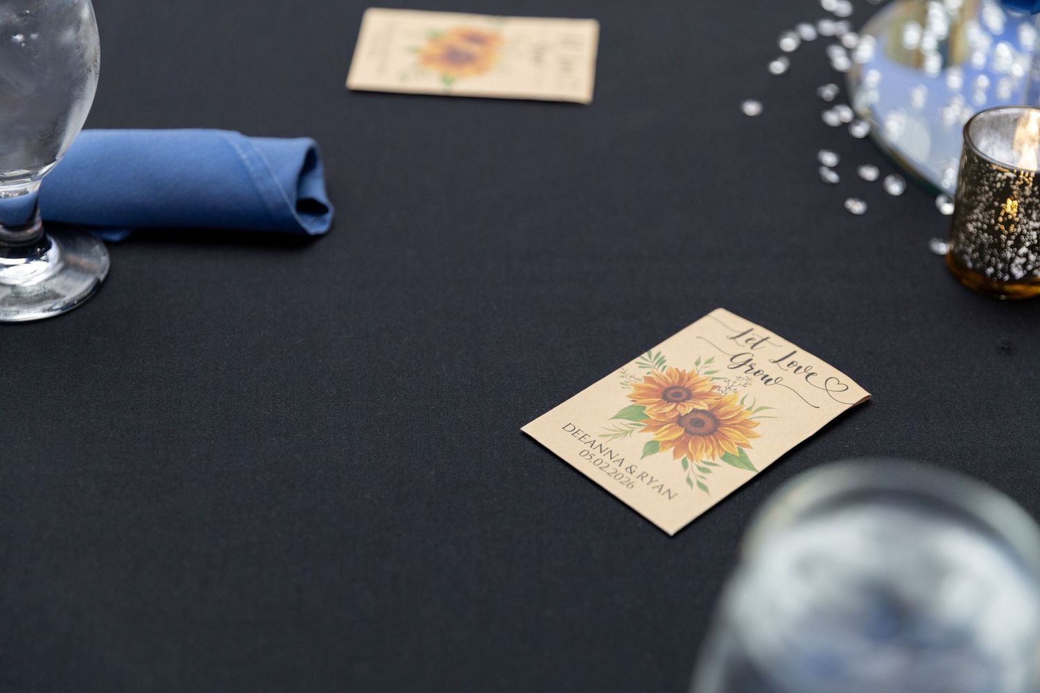 Sunflower wedding place cards on black tablecloth with blue napkin, wine glasses, and decorative beads.