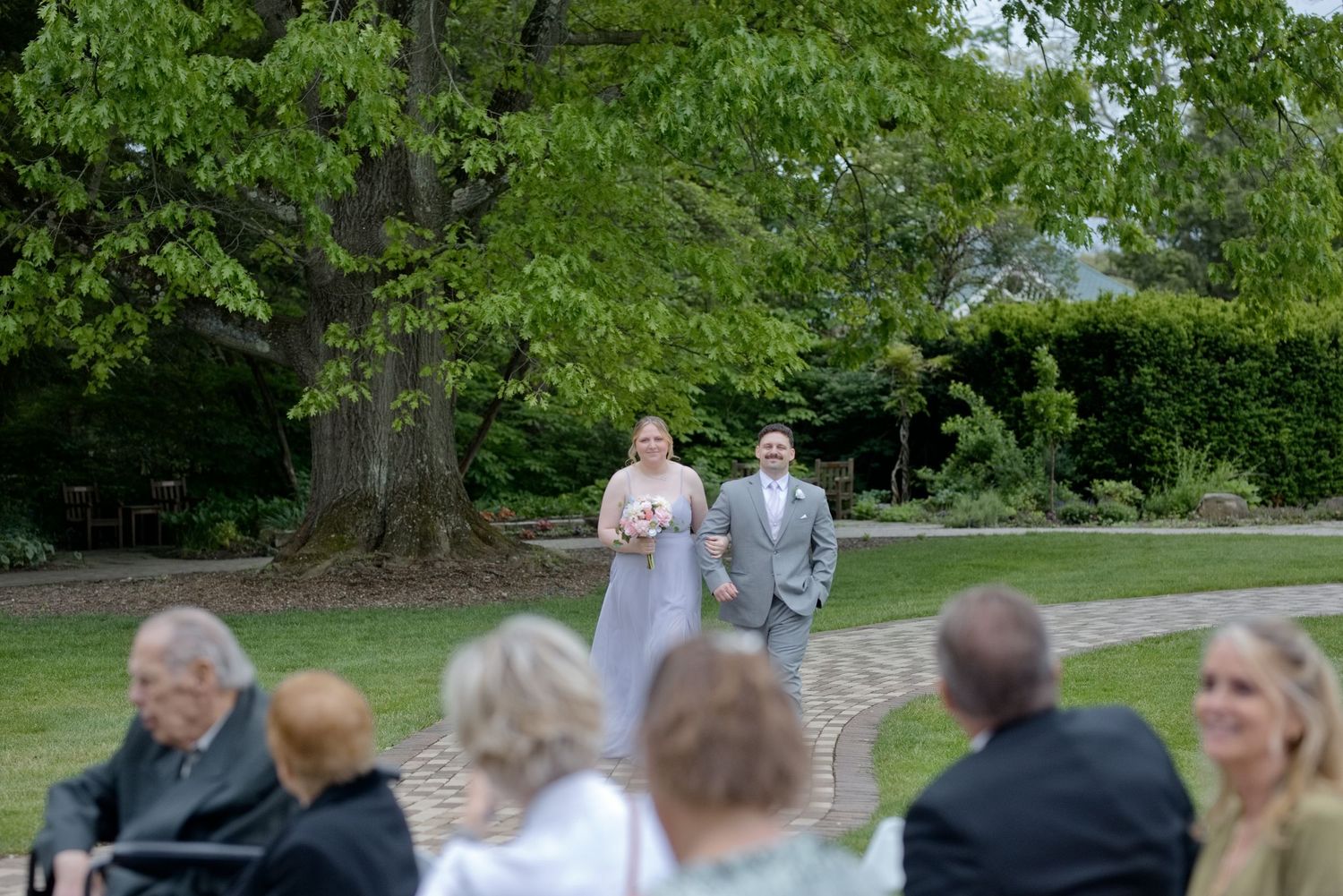 Bride and groom walking down outdoor garden wedding aisle surrounded by lush green trees and seated guests.