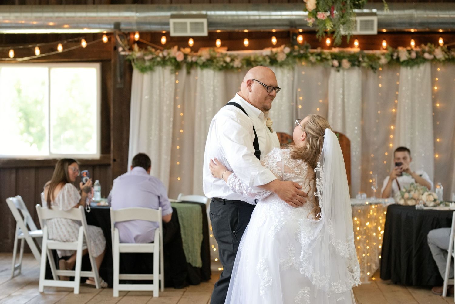 Bride and groom share their first dance at a rustic barn wedding reception decorated with string lights and greenery.
