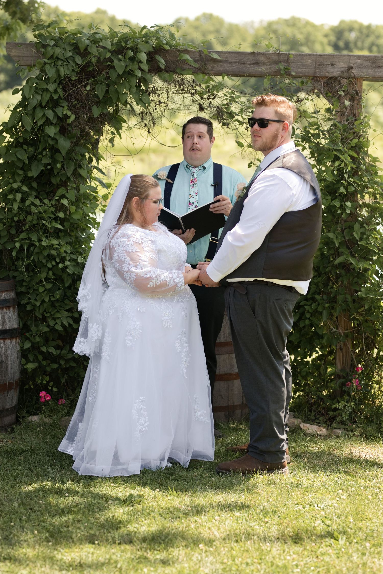 Outdoor wedding ceremony with bride in lace gown, groom in vest, and officiant reading vows under rustic wooden arch.