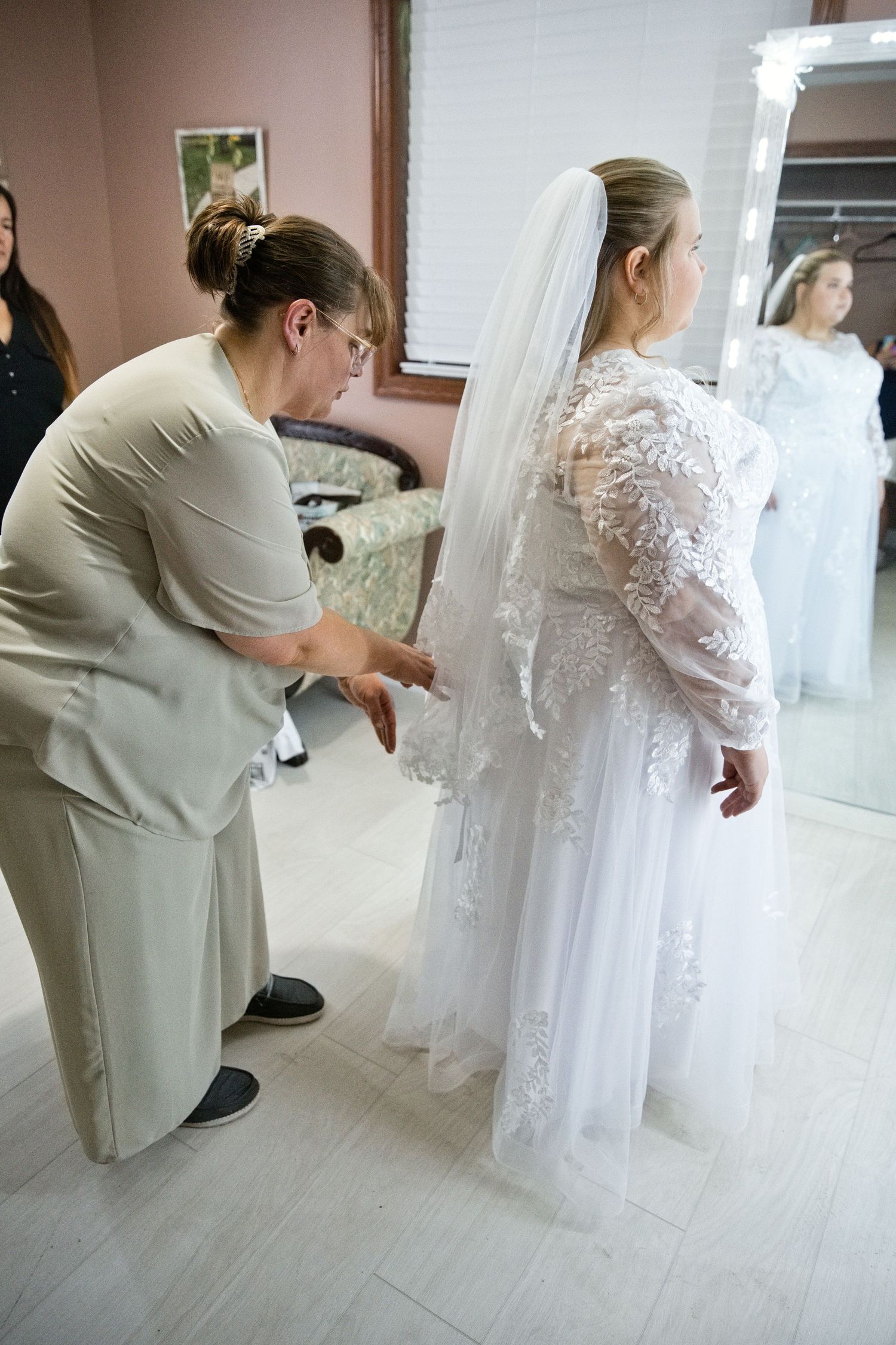 A bridal consultant helps a bride in a lace wedding gown and veil with final dress adjustments in a bridal boutique.