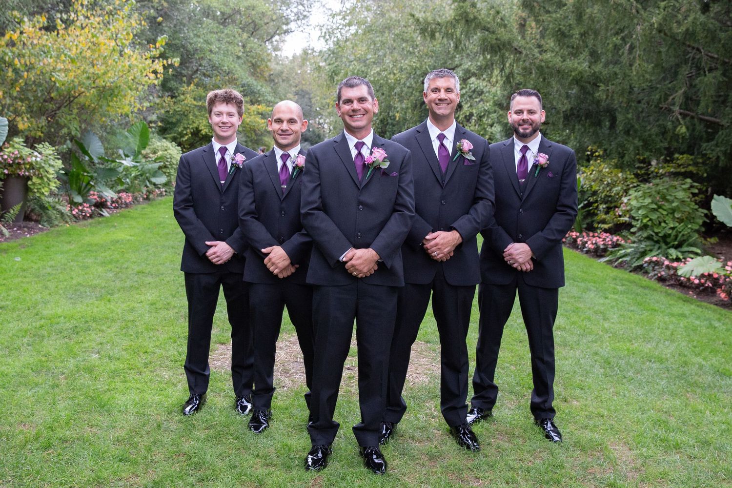 Groomsmen in formal attire lined up for photo in outdoor garden.