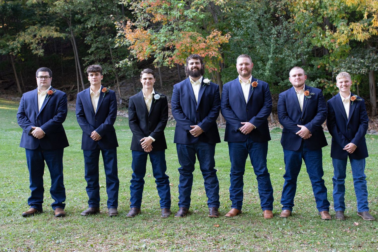 Groomsmen in navy suits standing in line outdoors against autumn trees.