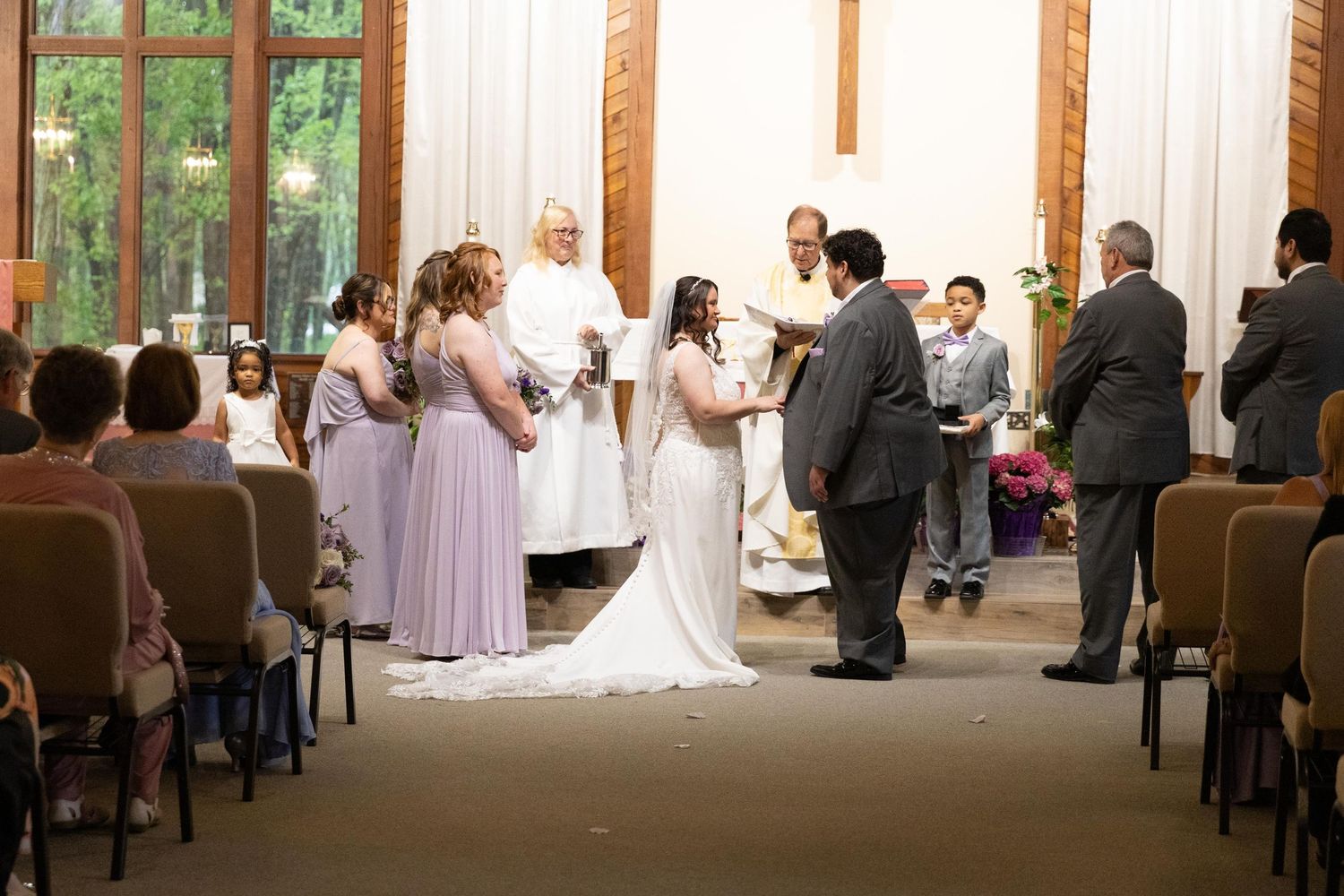 Bride and groom exchange vows at indoor wedding ceremony with bridesmaids, officiant, and guests in elegant chapel.