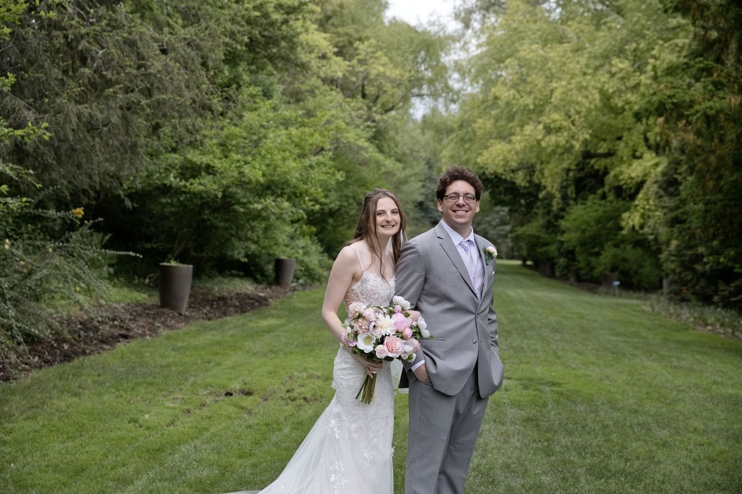 Bride and groom smiling outdoors on green lawn surrounded by lush trees on their wedding day.