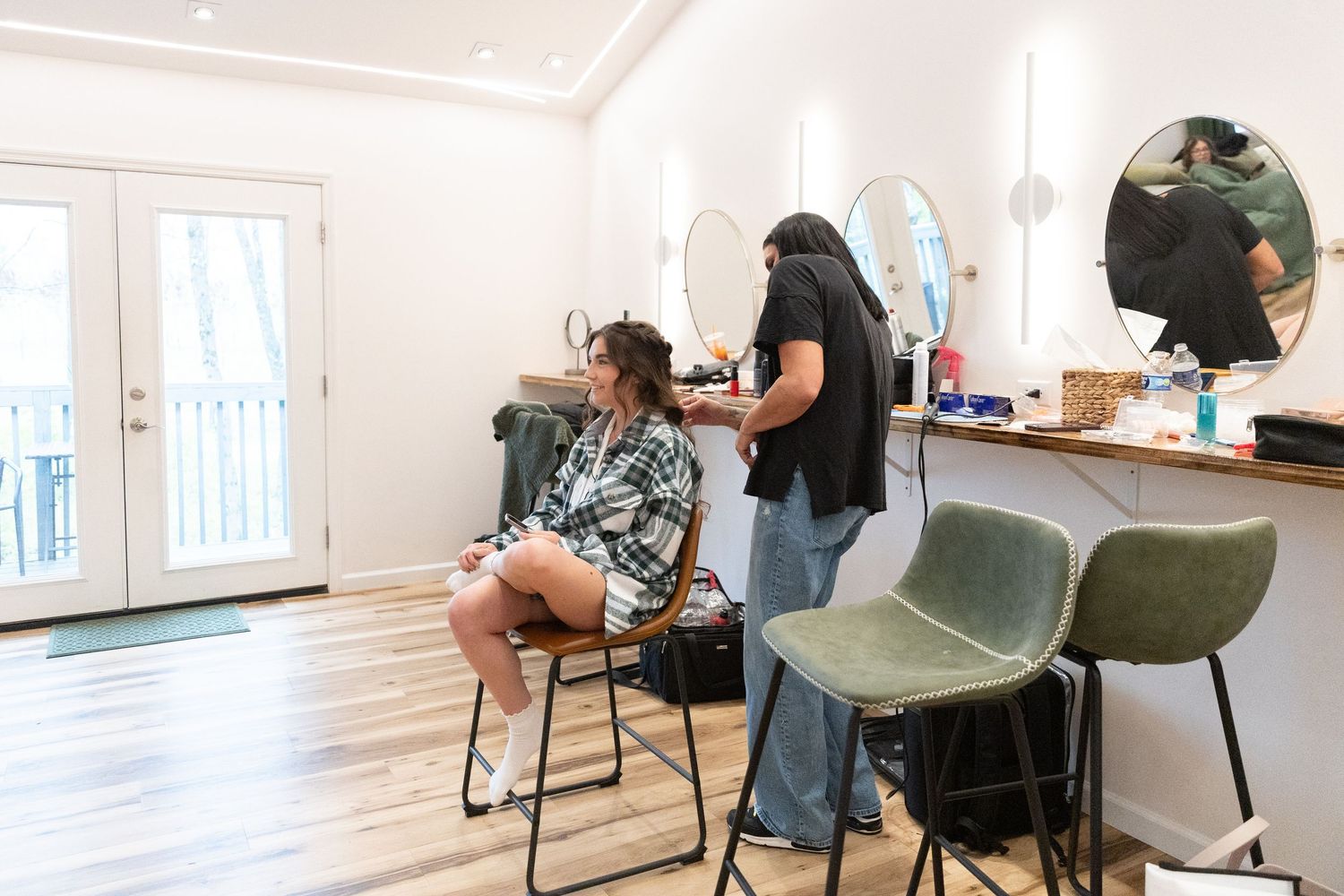 Hairstylist styling a client's hair in a modern salon with mirrors, green chairs, and bright natural lighting.