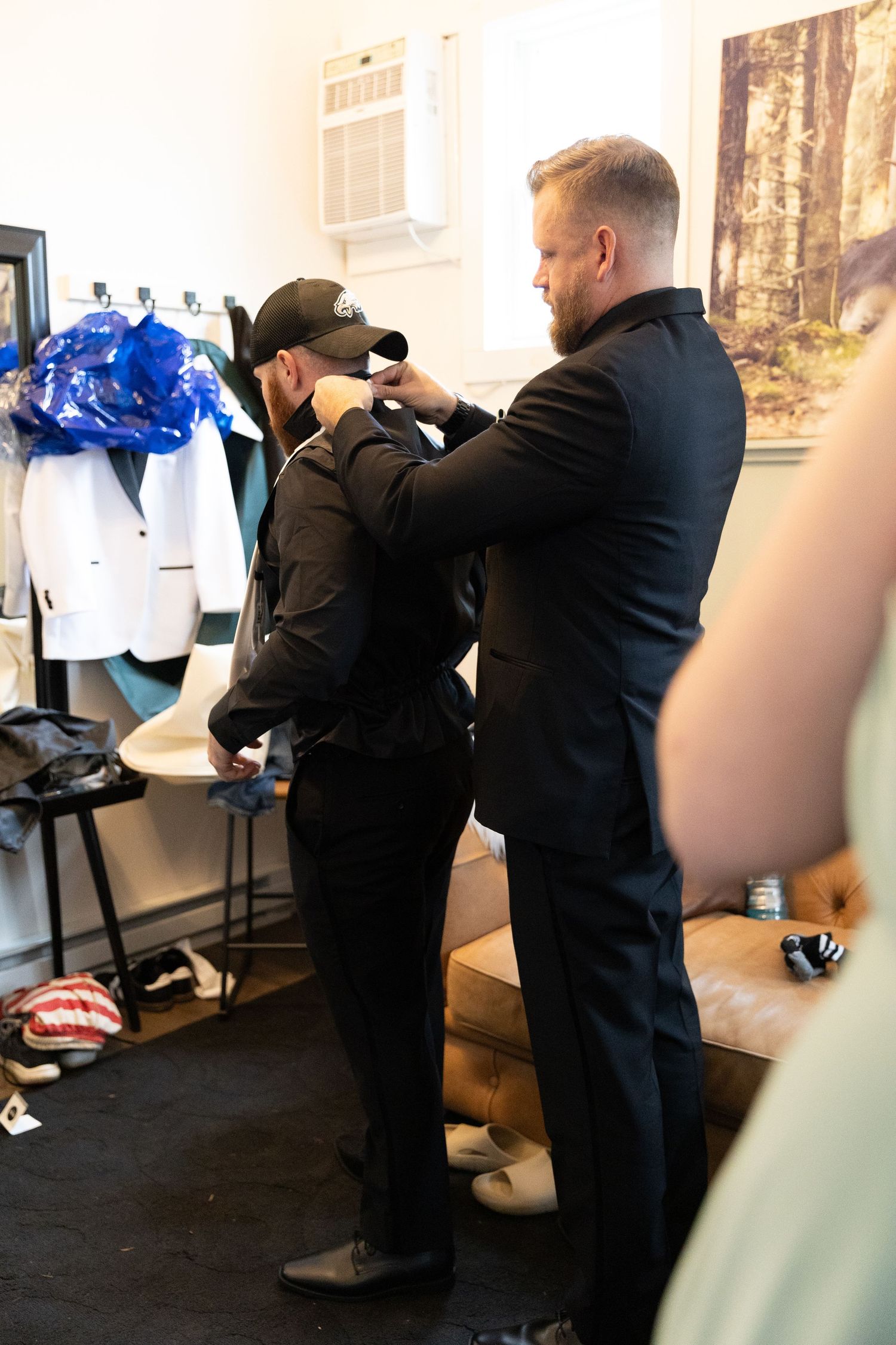 Two men in black suits in a dressing room, one helping the other adjust their collar before a formal event.