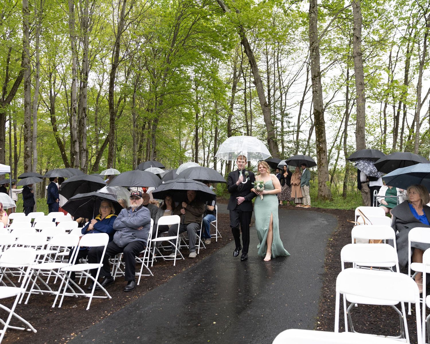 Outdoor wedding ceremony on a rainy day with guests holding black umbrellas seated in white chairs along a tree-lined aisle.