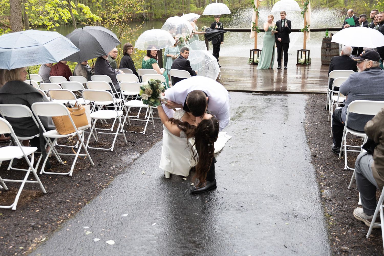 Bride and groom share a romantic kiss in the rain-soaked aisle during an outdoor wedding ceremony with guests holding umbrellas.