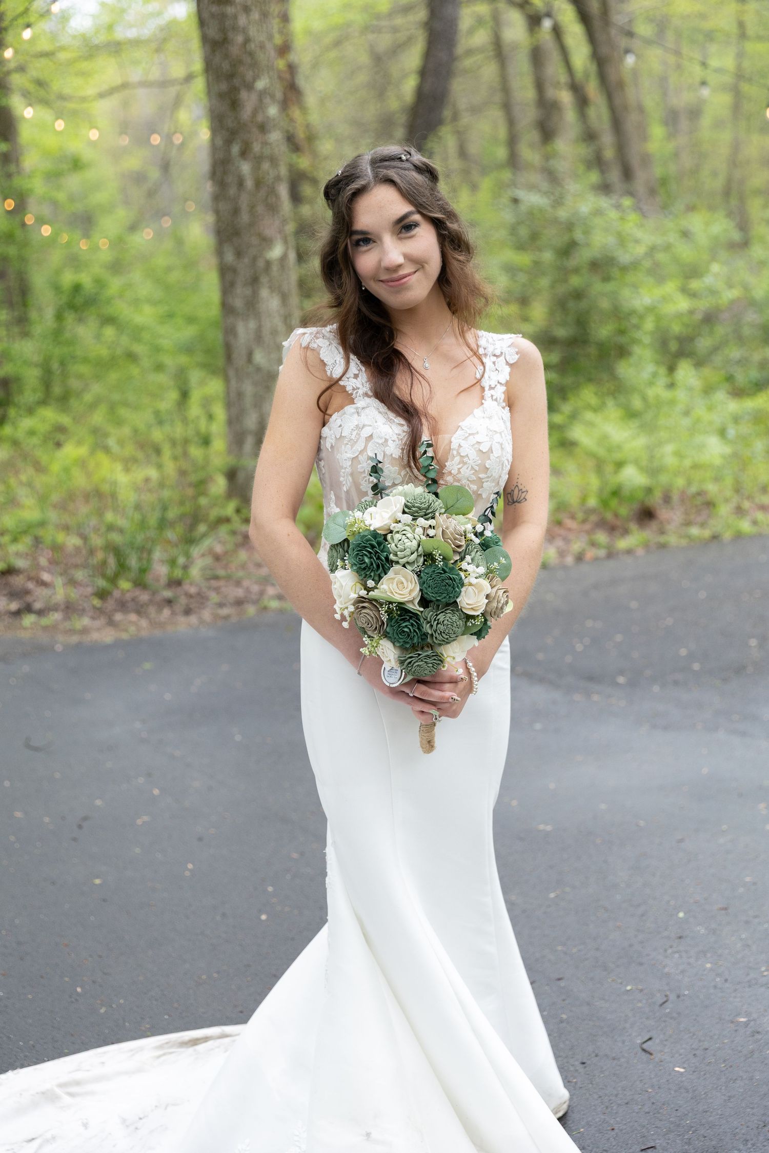Bride in white lace mermaid gown holding green and white bouquet, smiling outdoors on a tree-lined path.