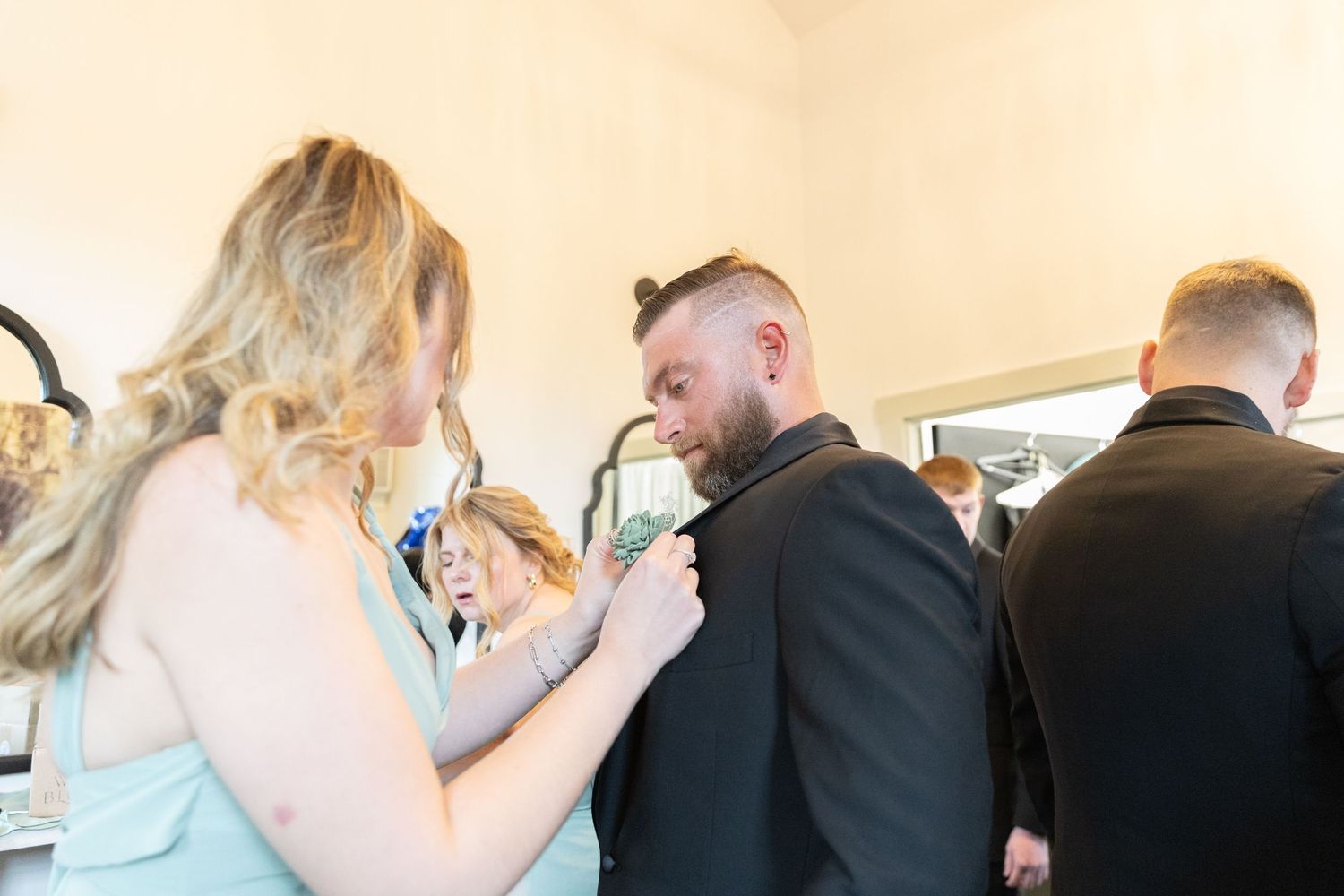 Woman in mint dress adjusting a bearded groom's suit jacket before a wedding ceremony.