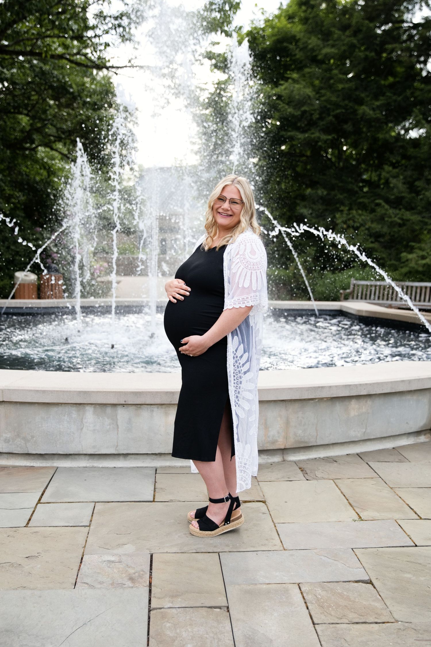 Pregnant woman in black dress and white cardigan poses by a stone fountain outdoors, cradling her baby bump.