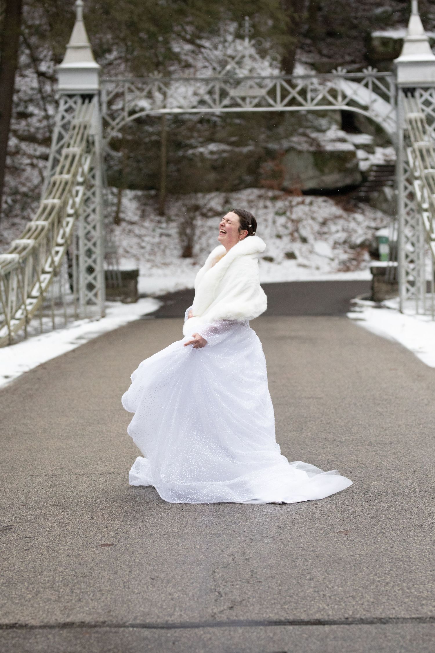 Joyful bride in white gown and fur wrap twirling on a snowy bridge, laughing with delight on her winter wedding day.