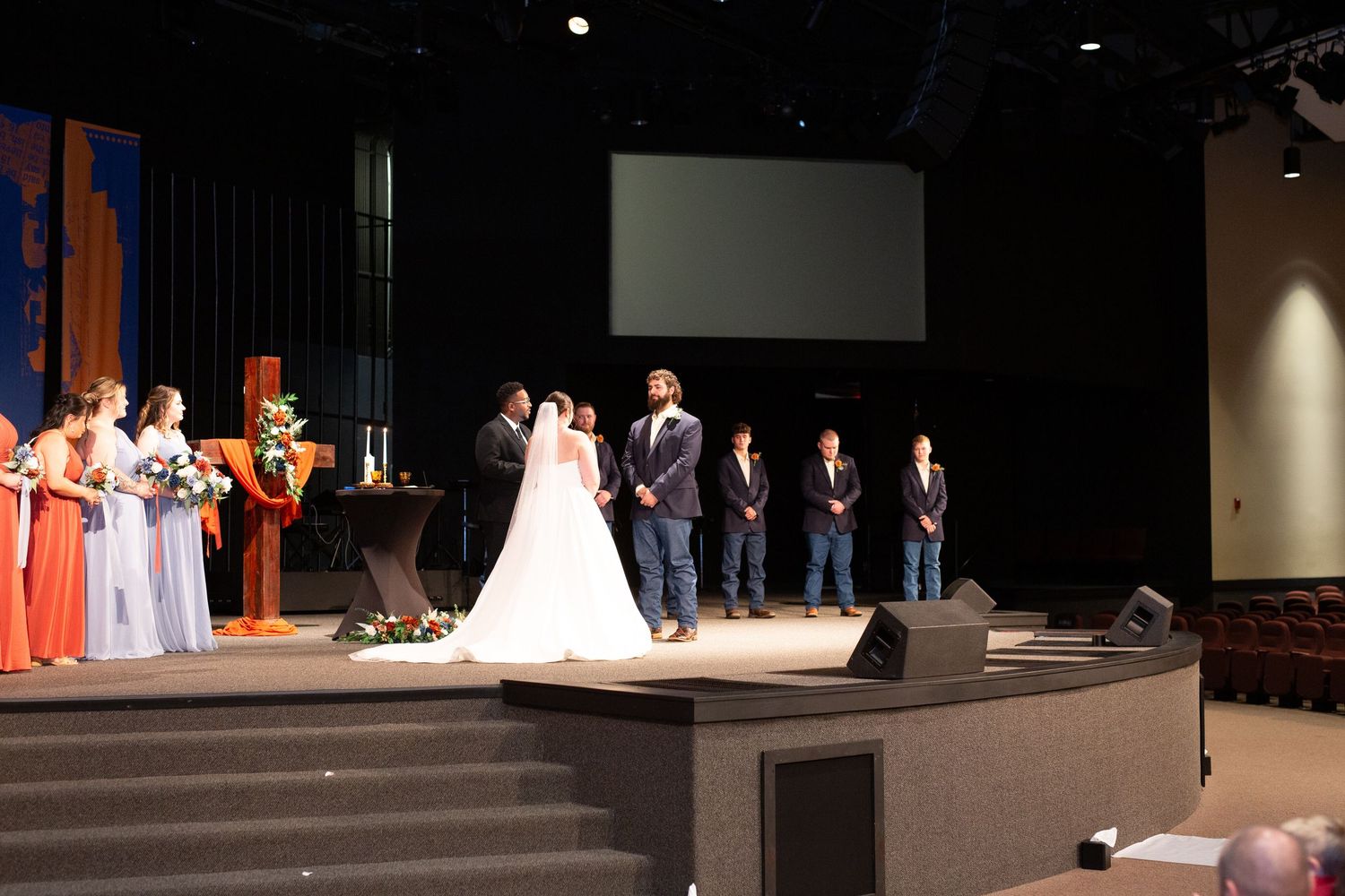 Wedding ceremony taking place on a stage with bridesmaids in orange and blue dresses and groomsmen in dark suits.