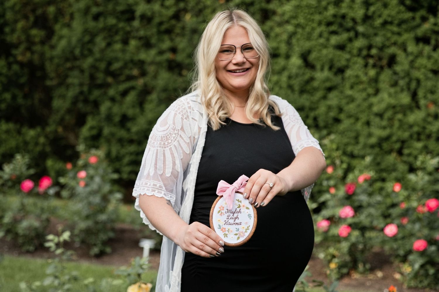 Pregnant woman in black dress holding embroidery hoop gender reveal sign outdoors near rose garden.