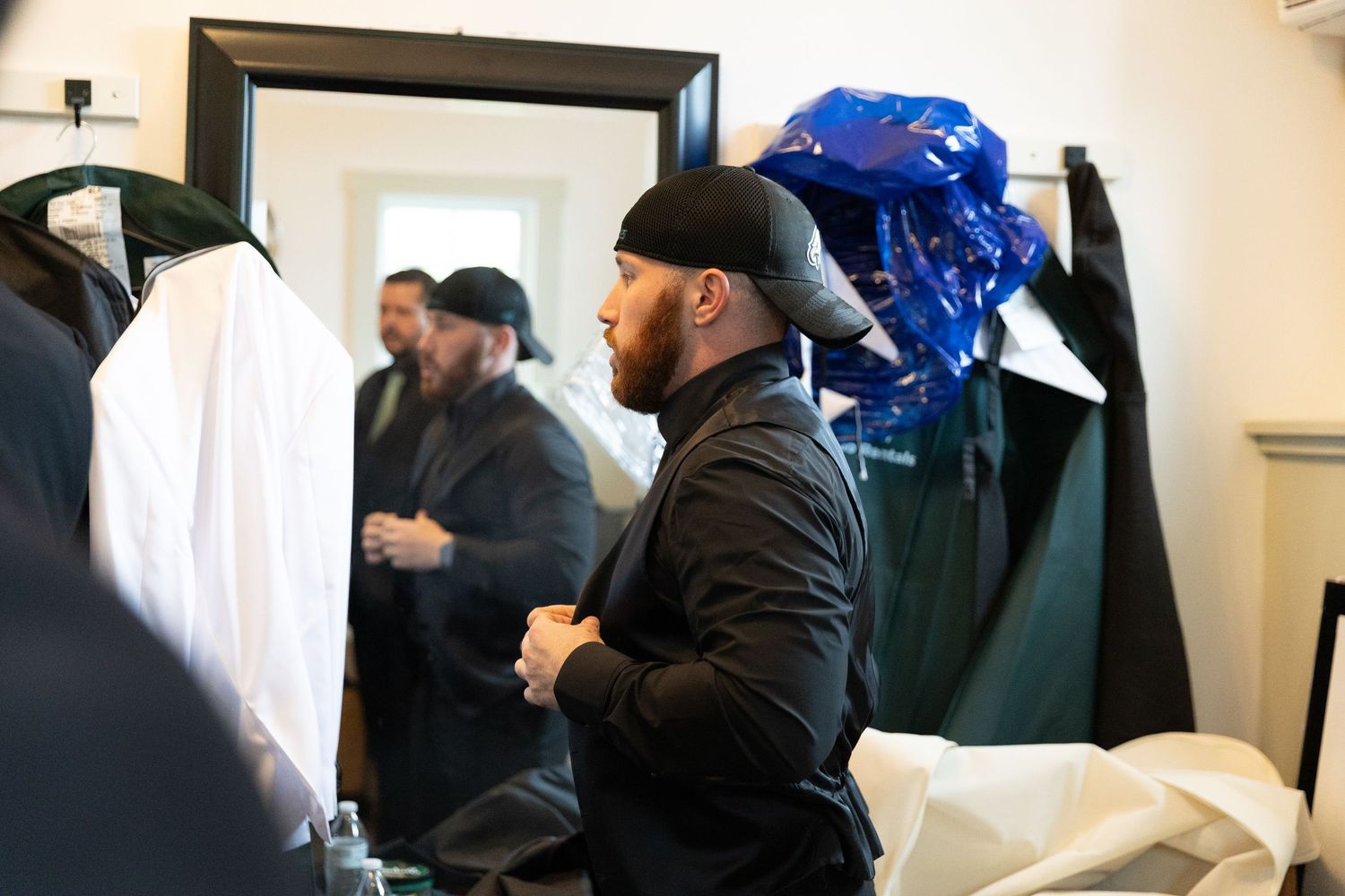 Man in black outfit and cap adjusting jacket in fitting room with mirror reflection and hanging garments.