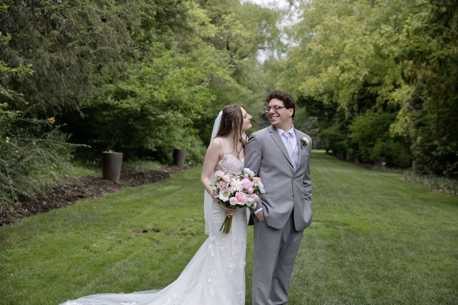 Bride holding pink bouquet and groom in gray suit share a joyful moment walking on a green lawn.