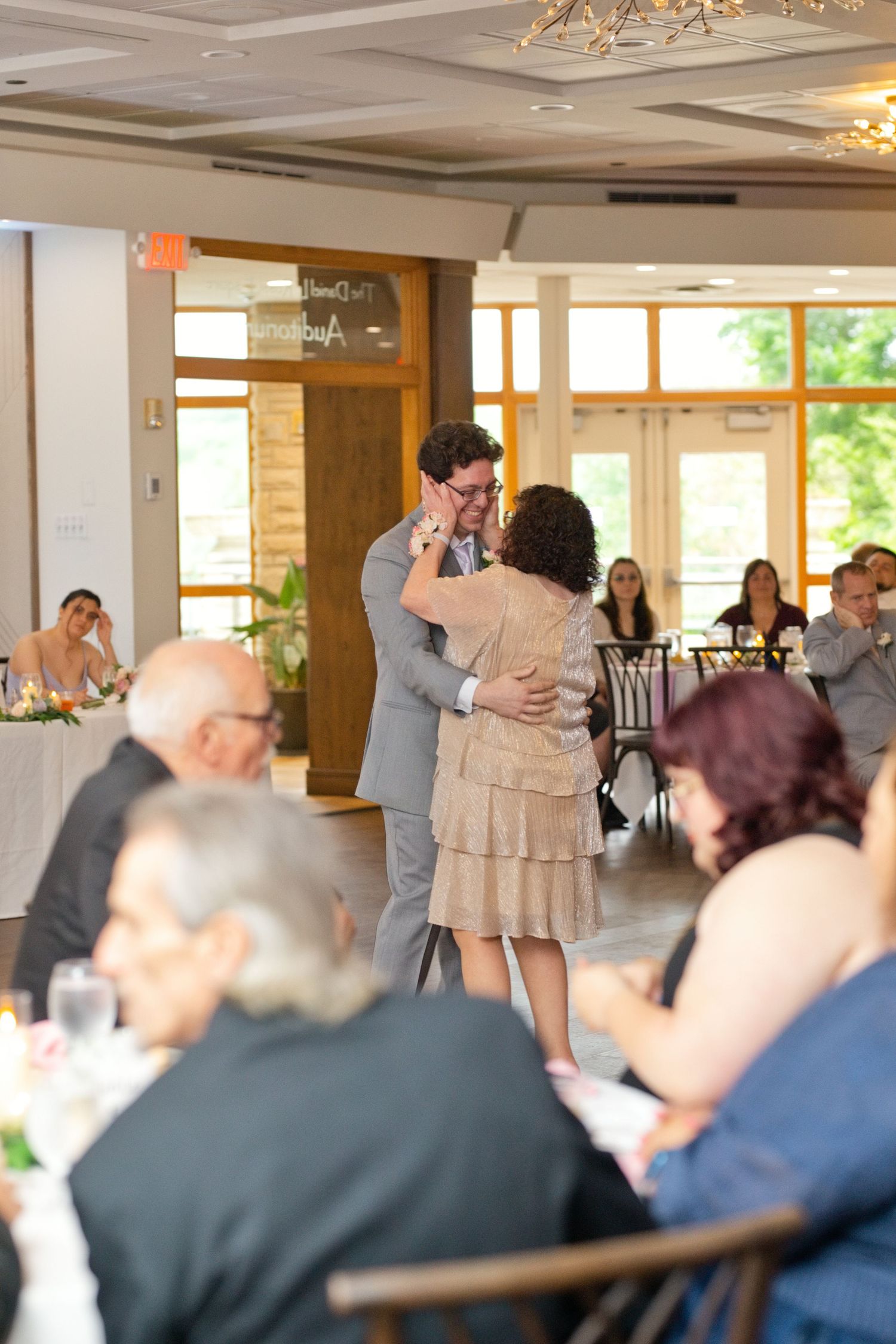 Groom shares an emotional mother-son dance at elegant wedding reception with guests seated at tables.