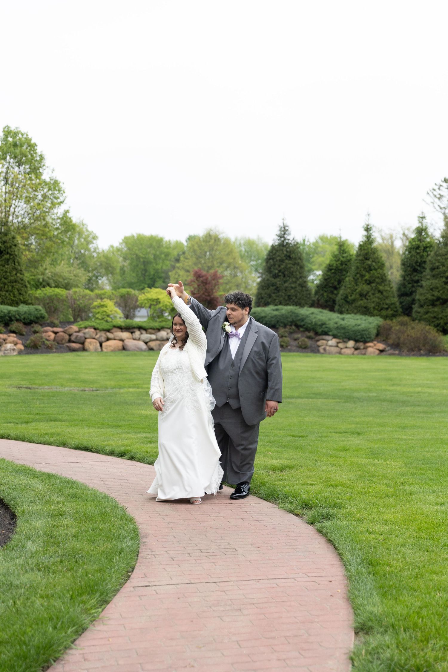 Bride and groom walk hand-in-hand on a brick path in a lush green garden, groom spinning bride in white gown.