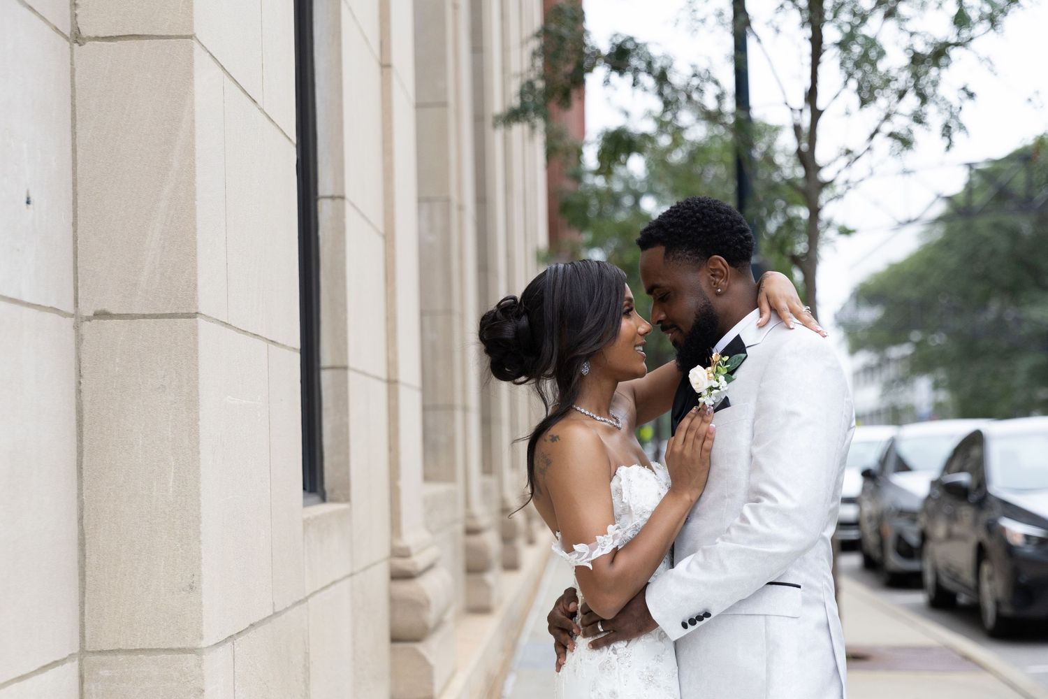 A Black bride and groom in white attire share an intimate moment outside a stone building on their wedding day.