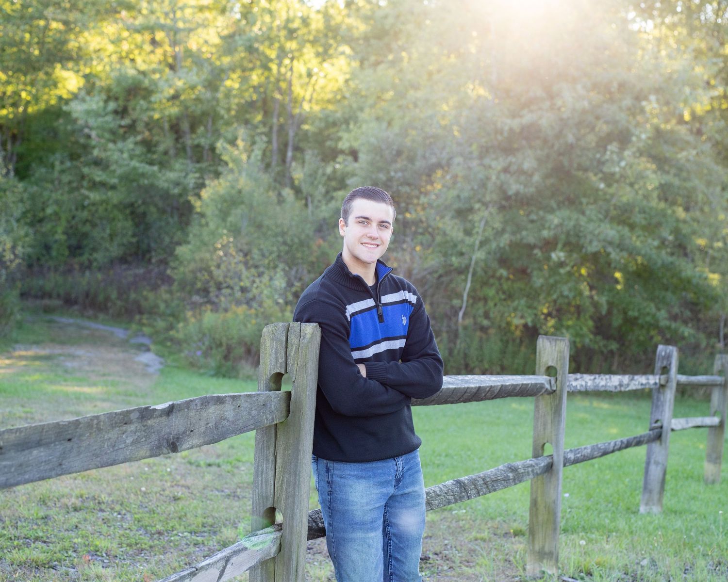Young man in a black and blue pullover leans on a wooden fence, smiling, with trees and sunlight in the background.