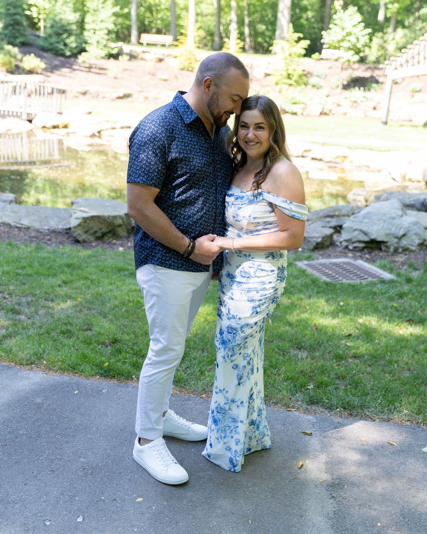 Couple posing together in a park, woman in floral dress, man in navy shirt and white pants.
