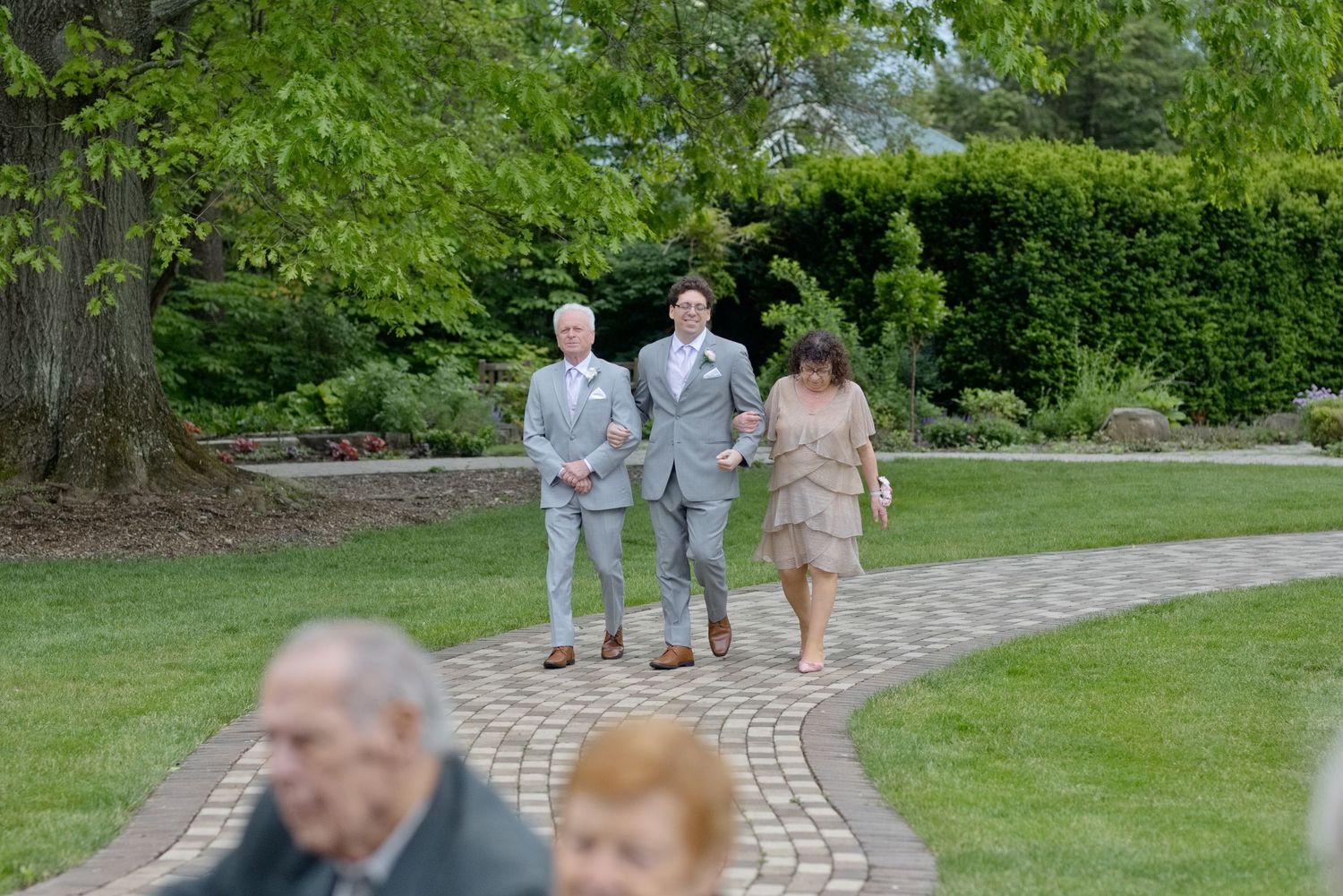 Groom walks with parents down brick garden path at outdoor wedding ceremony surrounded by lush greenery.
