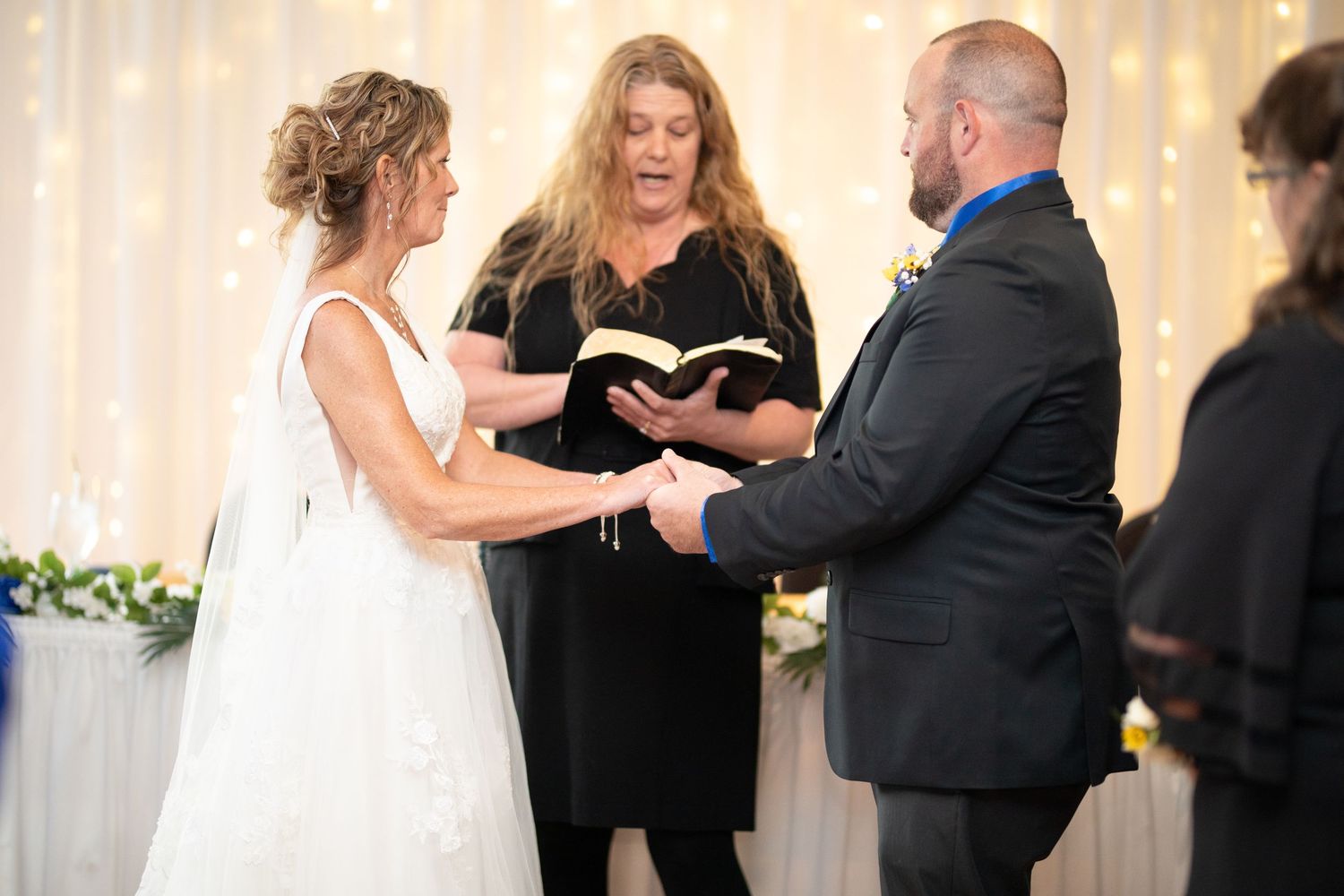 Bride and groom exchange vows at indoor wedding ceremony with officiant holding book, fairy lights backdrop.