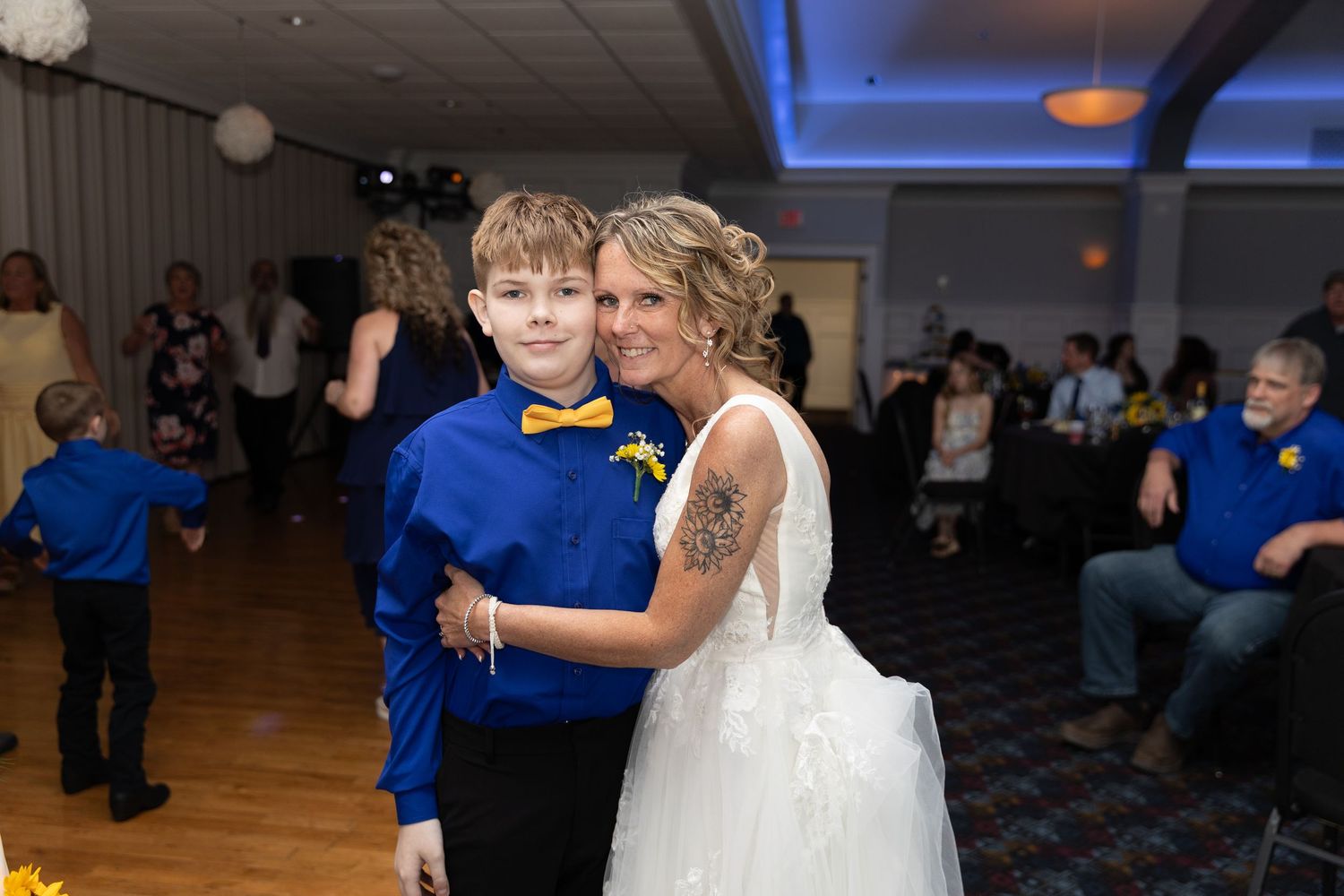 Bride in white dress hugging young boy in blue suit with yellow bow tie at wedding reception dance floor.