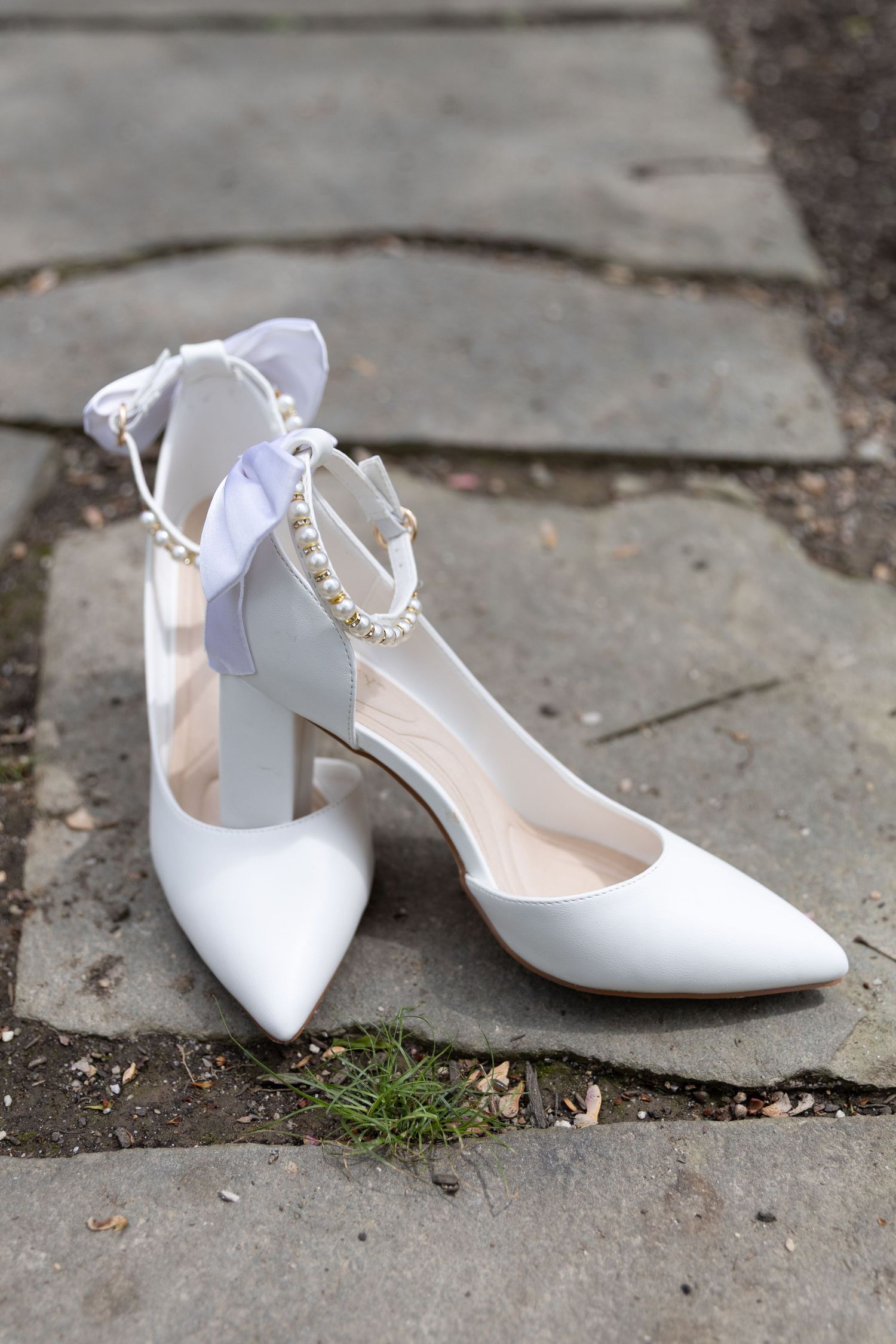White bridal heels with pearl ankle strap detail displayed on stone pavement, elegant pointed toe wedding shoes.