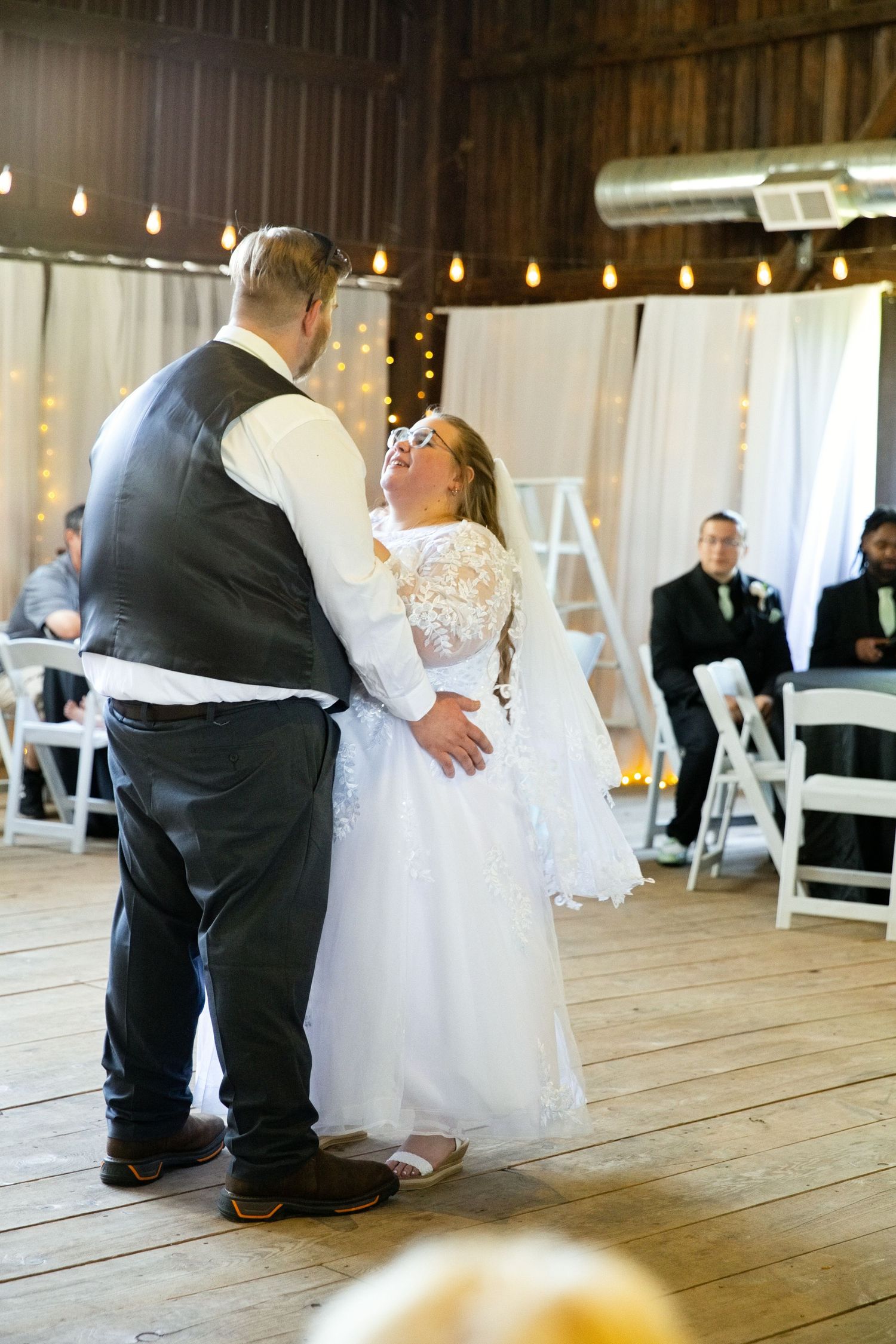 Bride and groom share their first dance in a rustic barn wedding venue decorated with string lights and white draping.