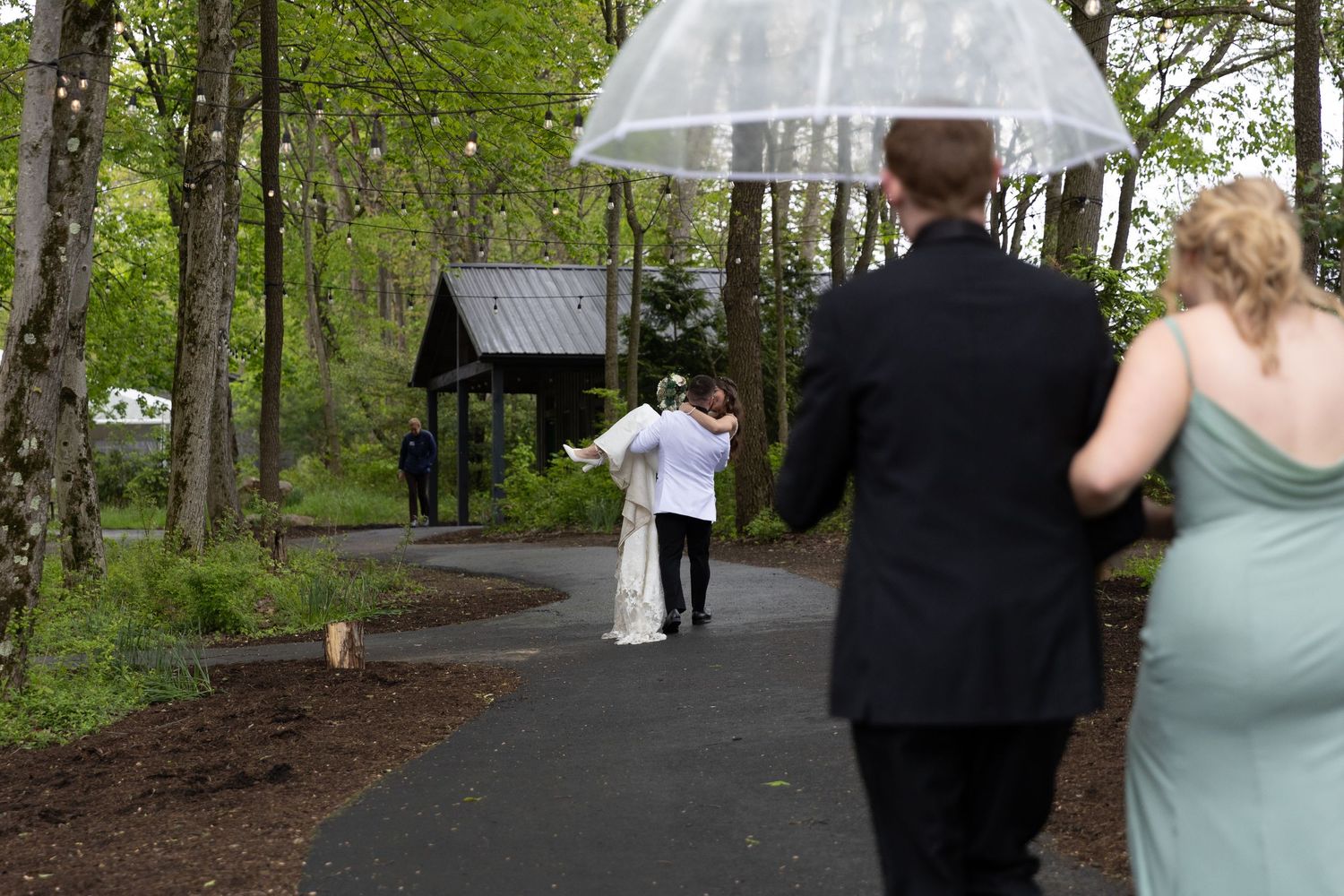 Wedding party walks along a wooded path toward a gazebo, with a couple holding a clear umbrella in the foreground.