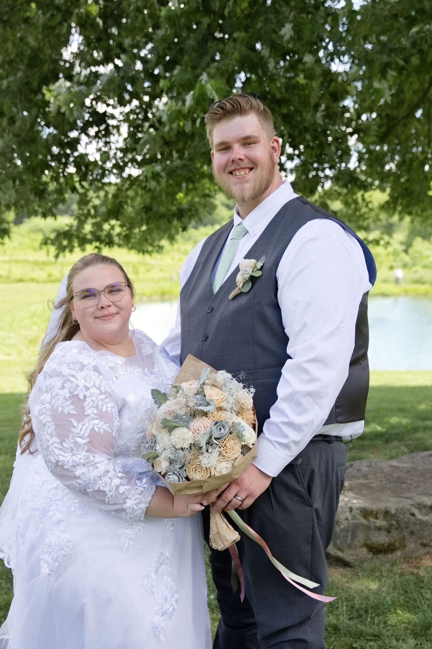 Bride and groom posing outdoors near a pond, bride holding a bouquet of neutral-toned flowers in a lace wedding dress.