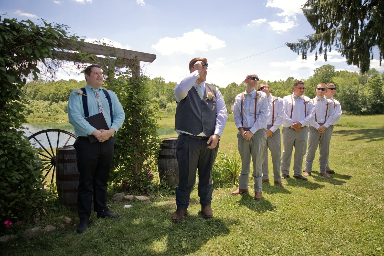Groomsmen in suspenders and ties stand outdoors on green lawn near rustic barrel and wooden arbor on sunny wedding day.