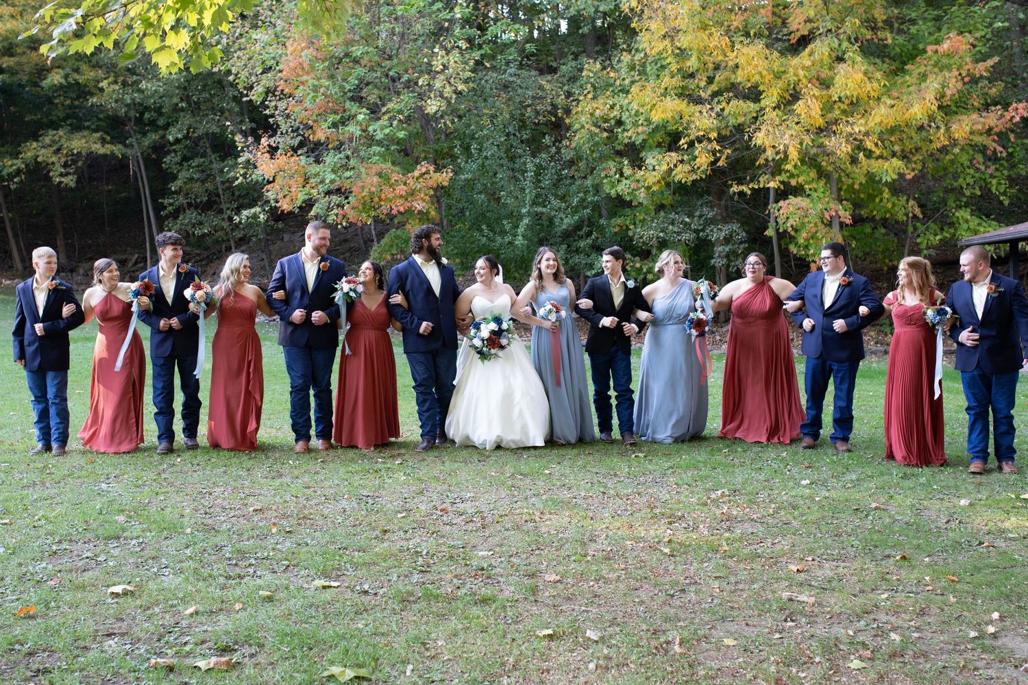 Wedding party standing in a line on grass with fall foliage backdrop.