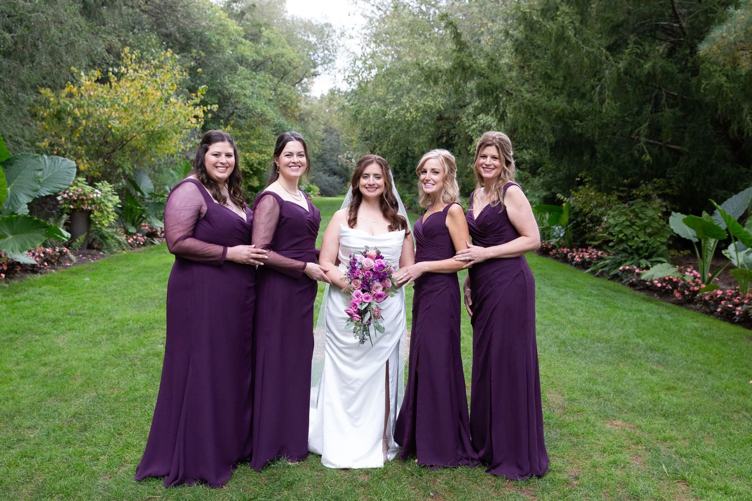 Bridal party in purple dresses posing together on green lawn.