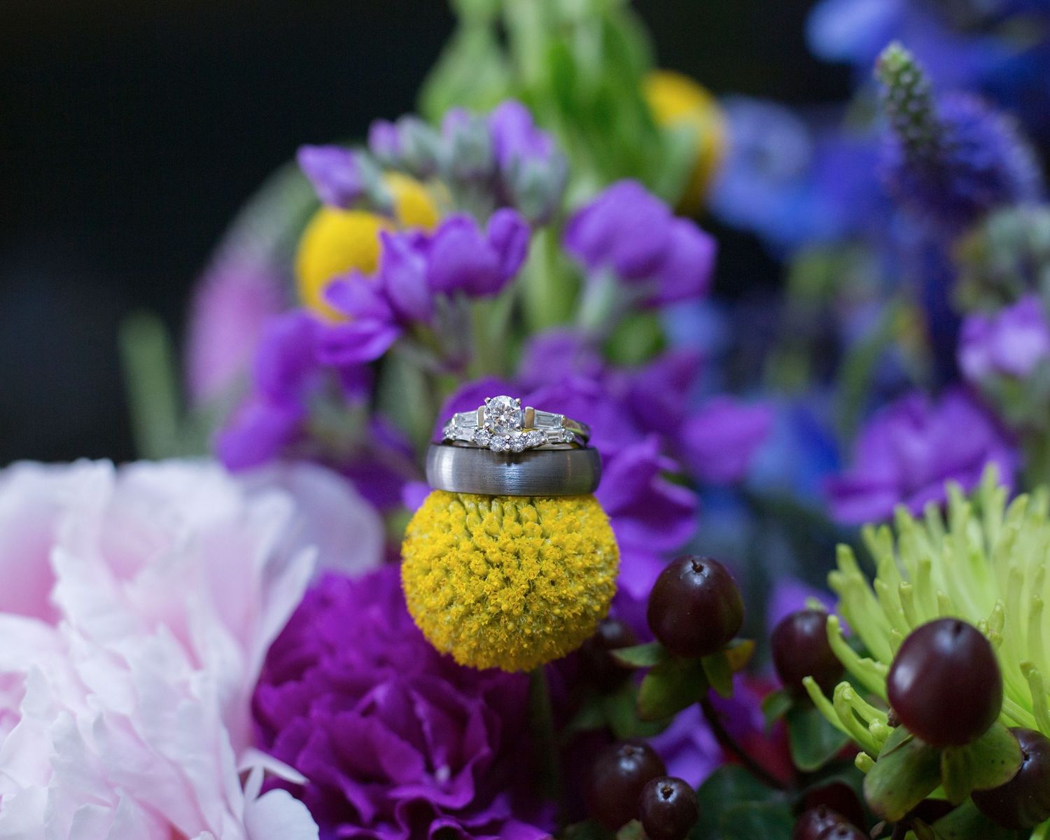 Wedding ring perched atop a yellow billy ball flower with purple blooms in the background.