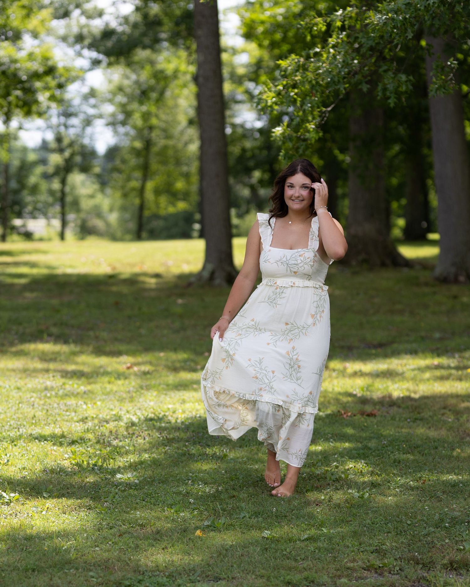 Woman in white floral midi dress walking barefoot through lush green park with tall trees in background.