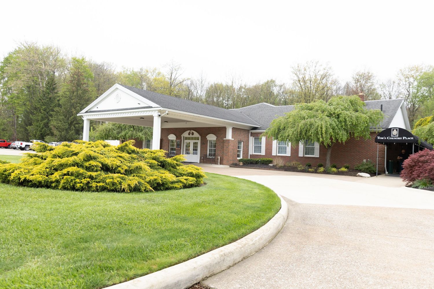 Elegant brick building with a circular driveway, manicured lawn, and blooming yellow shrubs surrounded by lush trees.