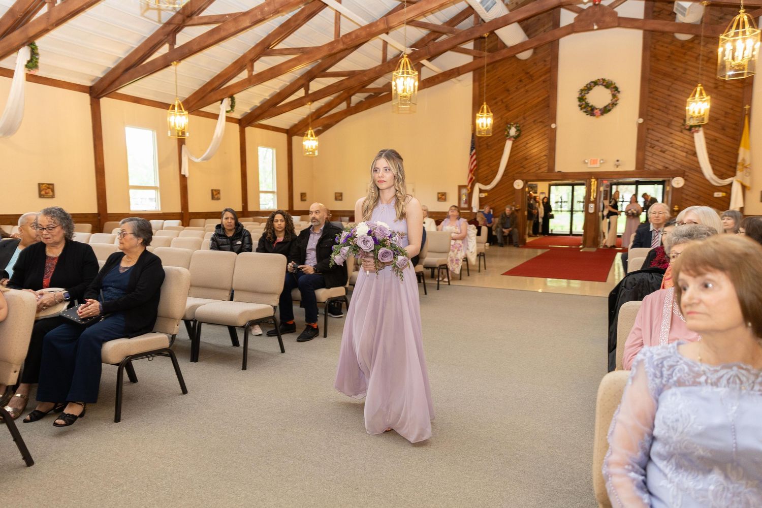 Bridesmaid in pink gown walks down church aisle during wedding ceremony with seated guests in wooden chapel.